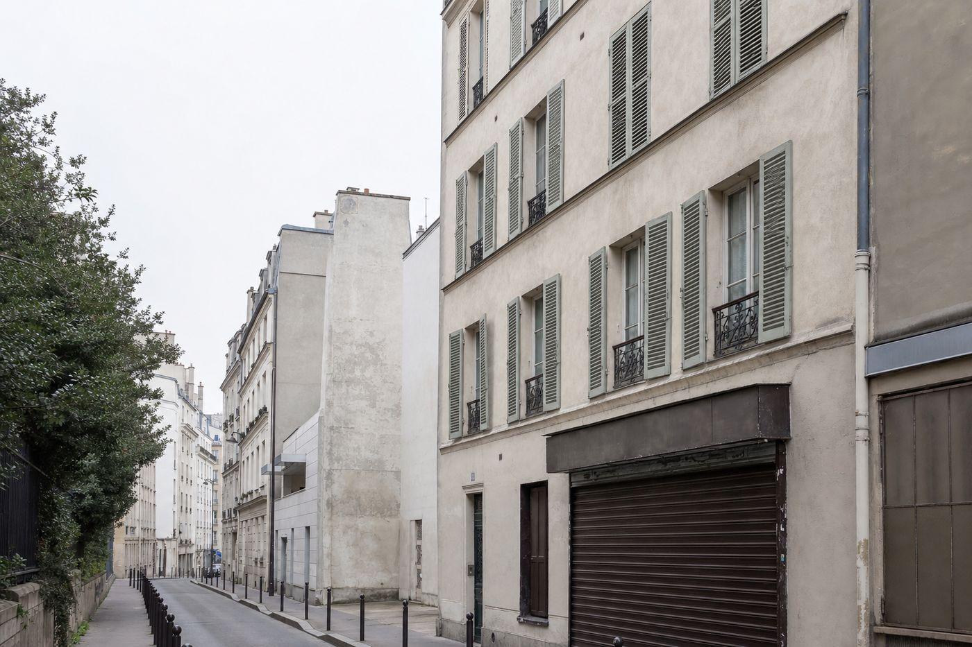 Rue étroite des Batignolles à Paris : immeuble de cinq étages en enduit clair, plus modeste qu’un immeuble haussmannien, persiennes métalliques fines vert-de-gris passées, boutique en rez-de-chaussée