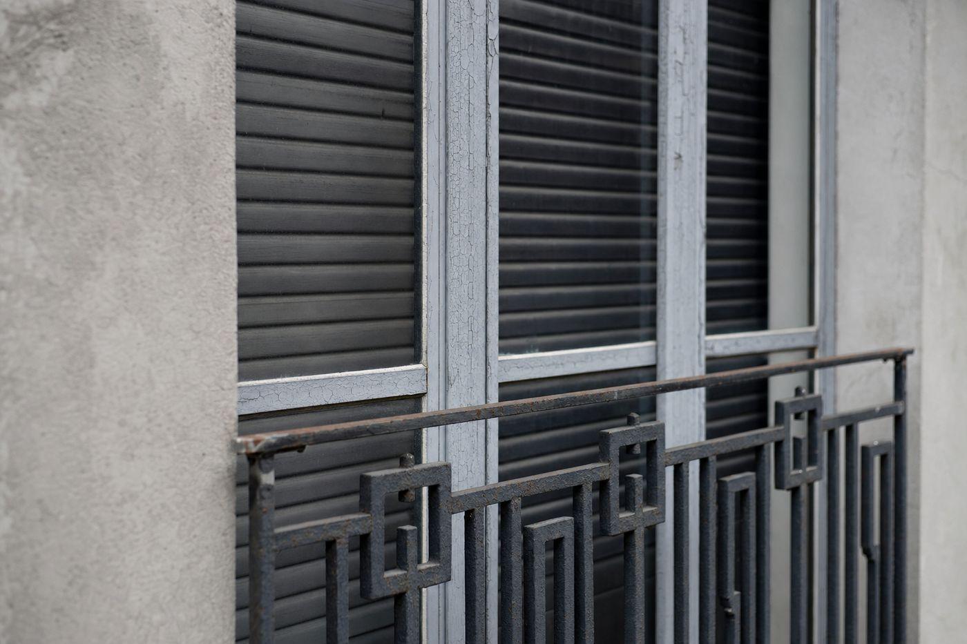 Détail d’une menuiserie en acier des années trente sur façade en béton enduit : profilés fins peints, garde-corps Art déco géométrique et volet métallique gris à lames étroites replié dans le tableau