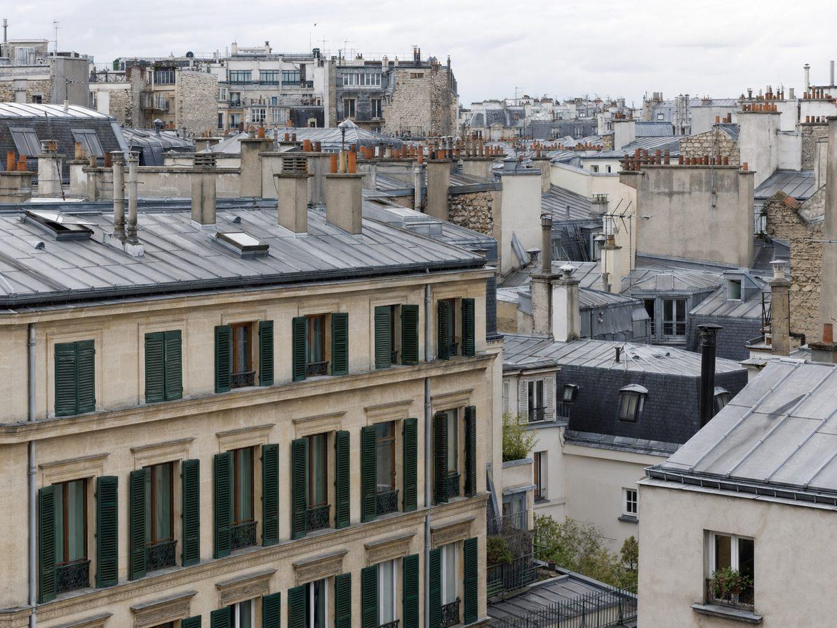 Vue en surplomb des toits de zinc parisiens et des étages supérieurs d'immeubles en pierre dont les fenêtres portent des persiennes métalliques vert foncé