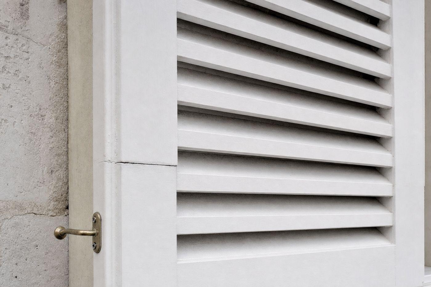 Volet bois persienné peint en blanc sur une façade en pierre claire, vu de biais : lames en enfilade, légère fissure à un assemblage, crochet en laiton maintenant le vantail ouvert