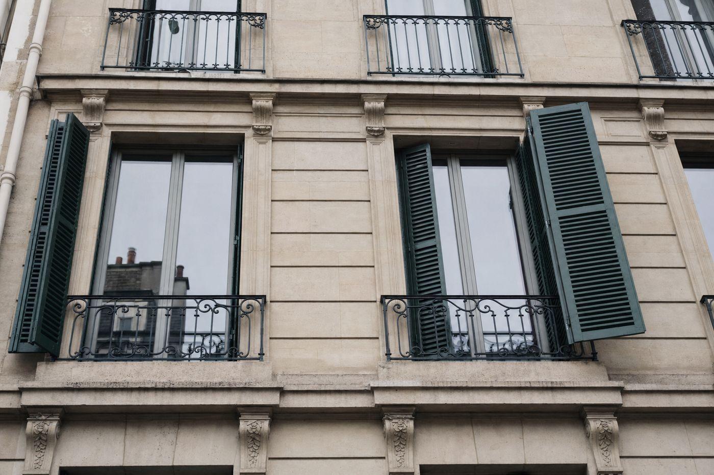 Baies très hautes d’un immeuble en pierre de taille de l’ouest parisien, balconnets en fer forgé et persienne métallique vert foncé repliée contre la pierre