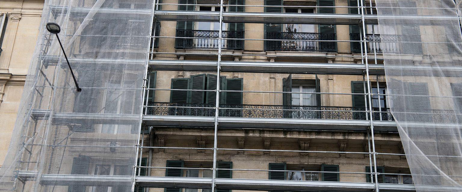 Immeuble haussmannien parisien en pierre de taille sous échafaudage, filet de protection écarté laissant voir quatre étages de persiennes vertes et de garde-corps en fer forgé