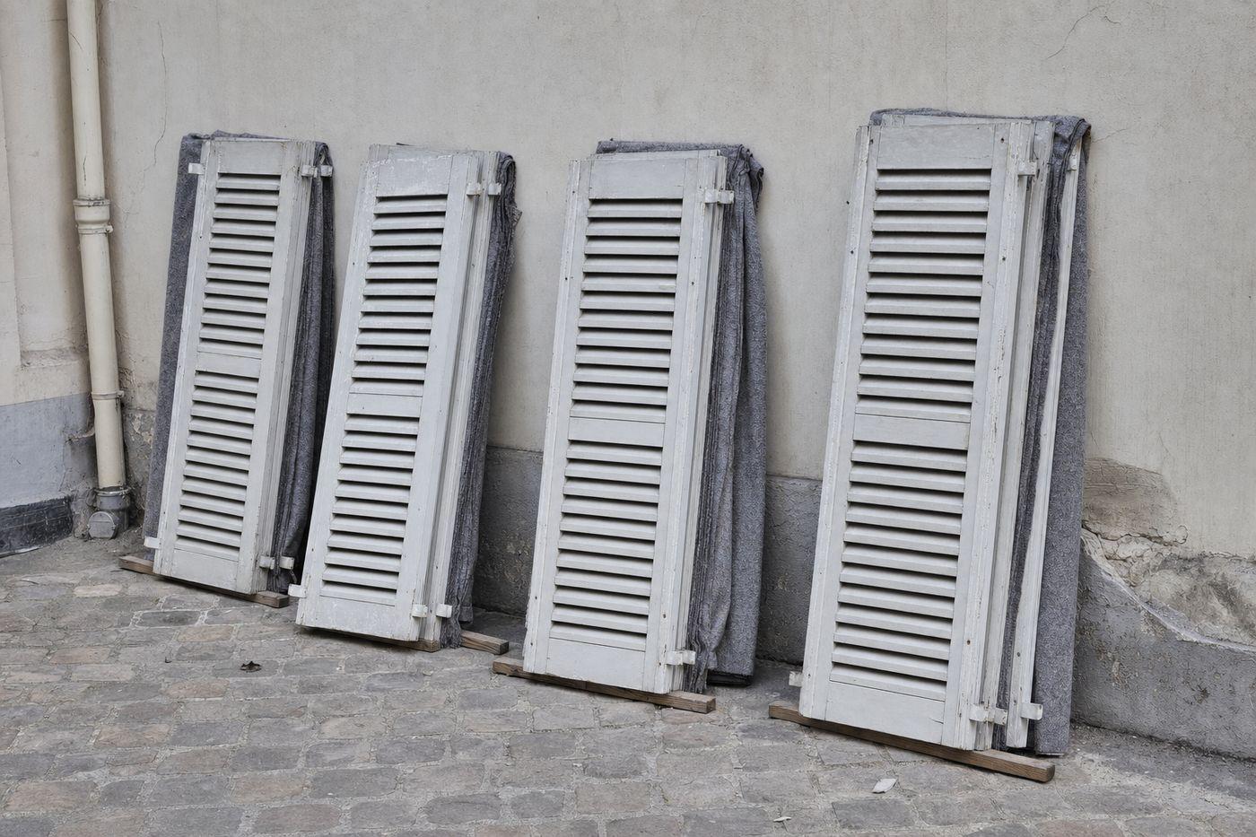 Vantaux de volets persiennés clairs déposés et rangés par paires contre le mur d'une cour pavée parisienne, calés sur des tasseaux et séparés par des couvertures de protection