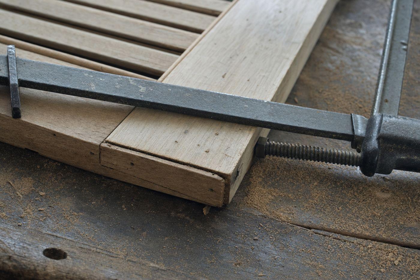 Angle inférieur d’un volet bois décapé posé sur un établi, assemblage à tenon et mortaise entre montant et traverse, jeu ouvert par le temps, serre-joint en place