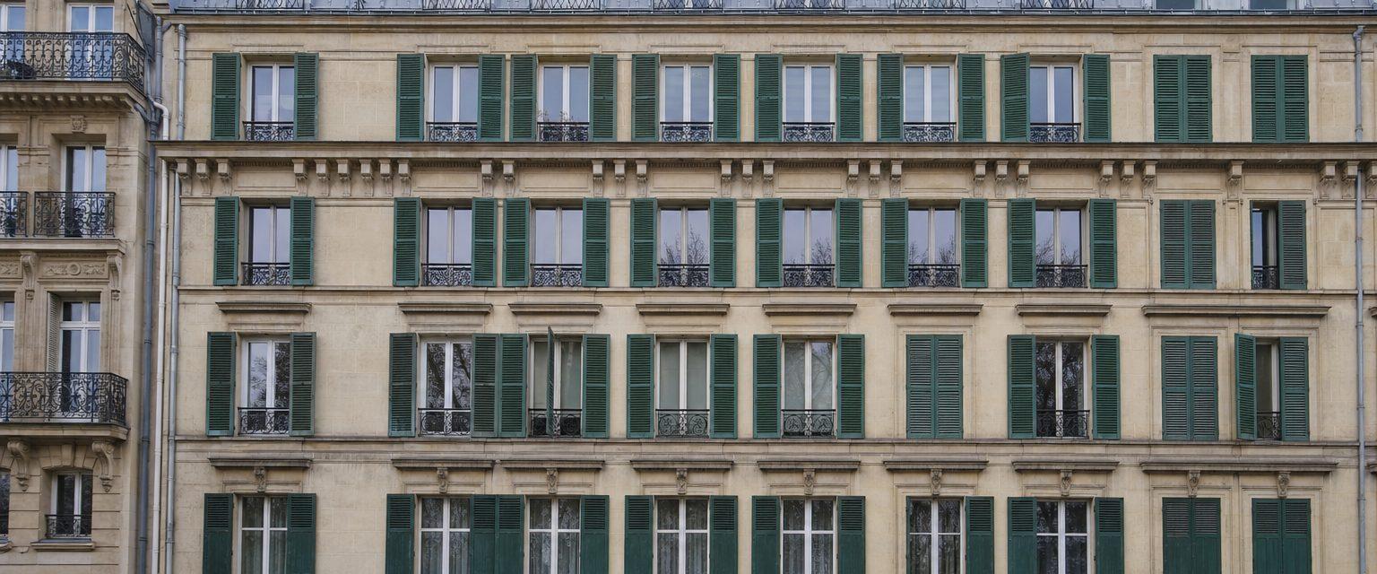Façade d'immeuble haussmannien parisien en pierre de taille, quatre étages de fenêtres à persiennes vertes et garde-corps en fer forgé